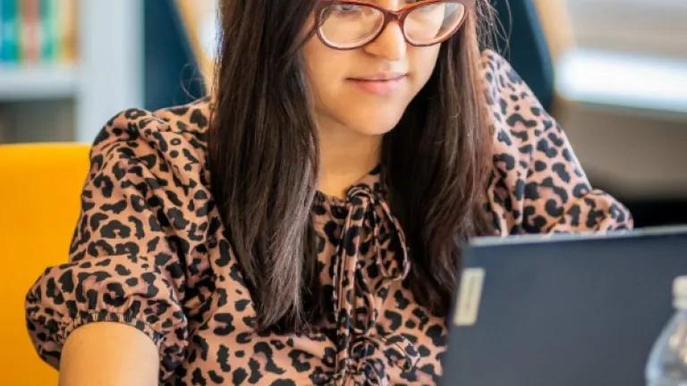 A woman seated at a desk working at a laptop. A bookshelf can be seen in the background.