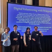 Three student and the project leader posing happily in front of the screen listing awards for Digital Volunteering with Wikipedia