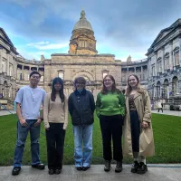 Five students standing in the Edinburgh Old College courtyard. Background: a lawn, encircling campus buildings, a tower