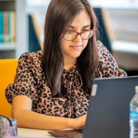 A woman seated at a desk working at a laptop. A bookshelf can be seen in the background.