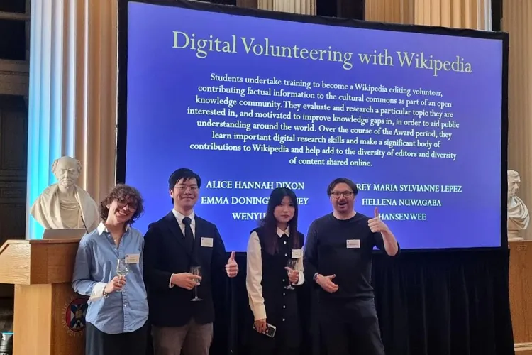 Three student and the project leader posing happily in front of the screen listing awards for Digital Volunteering with Wikipedia
