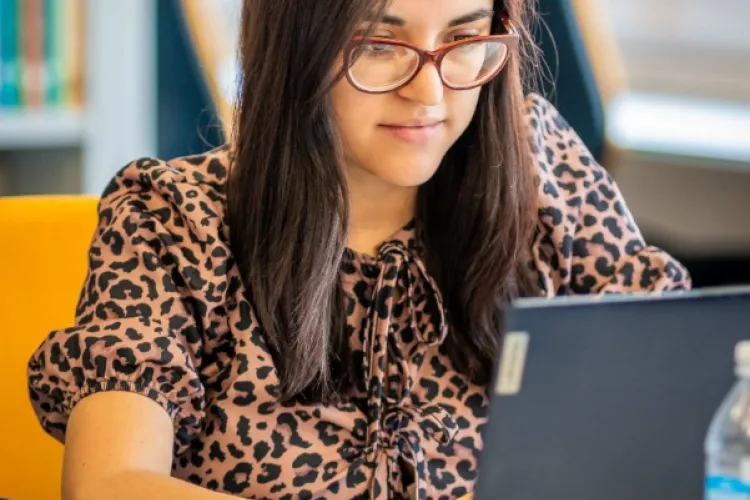 A woman seated at a desk working at a laptop. A bookshelf can be seen in the background.