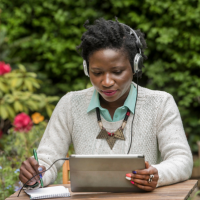 Woman sitting at laptop outside and studying, with a headset on her head. 