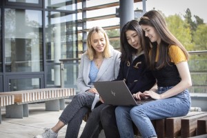 Three people sit outside a University building on a bench and look at a laptop.