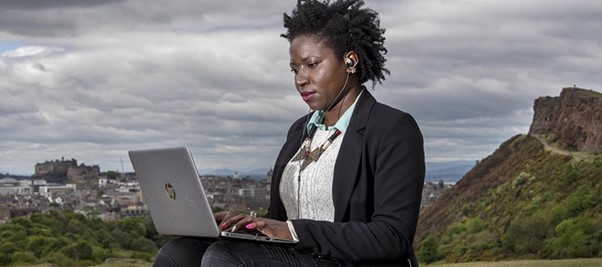 A woman sits in Holyrood Park, with Edinburgh Castle behind her to her left and Salisbury Crags her right, typing on a silver laptop which is placed on her lap.